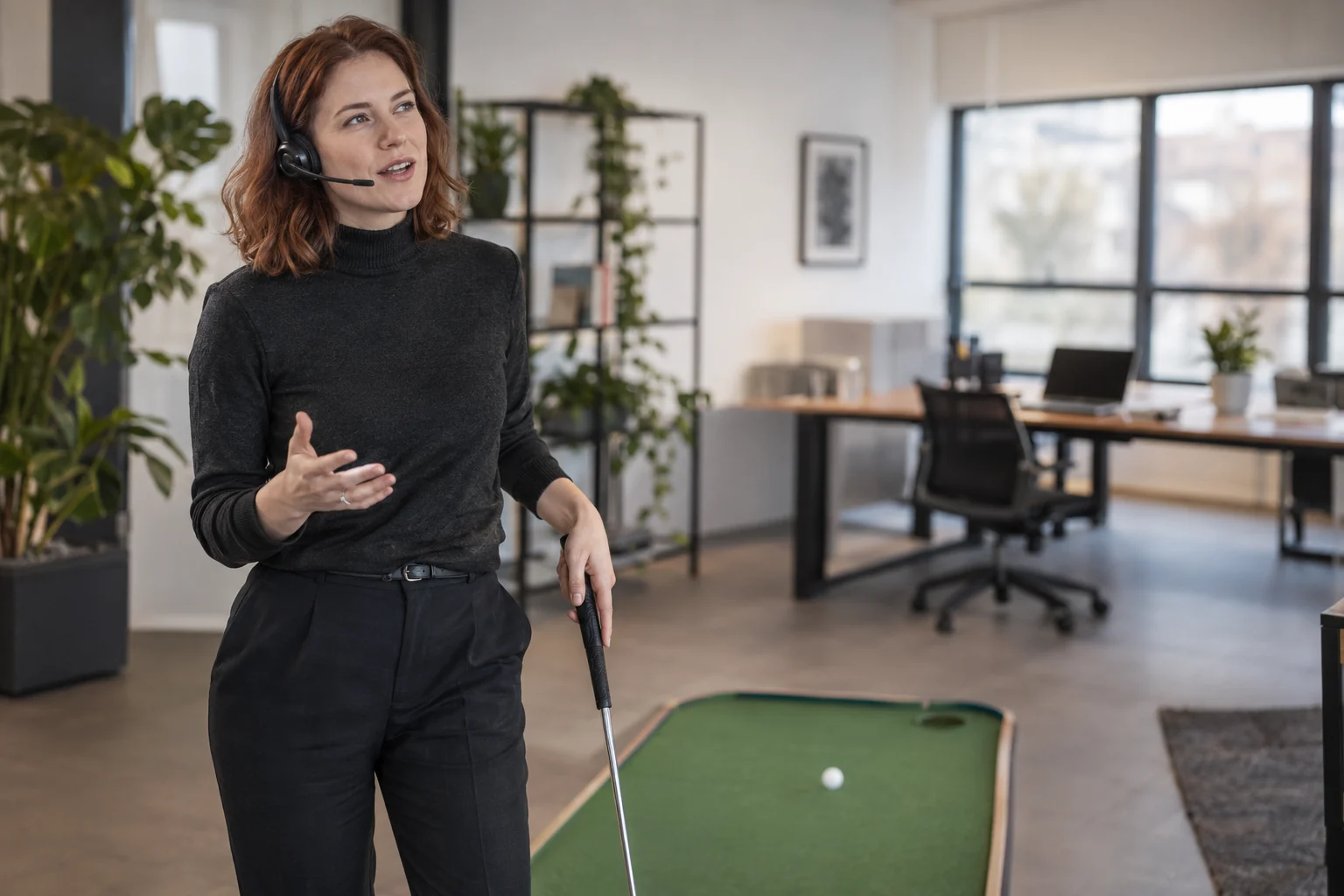 KI-generierte Szene: Eine Person hält beim Büro-Minigolf eine Idee über ihr Headset fest; der Laptop steht im Hintergrund.