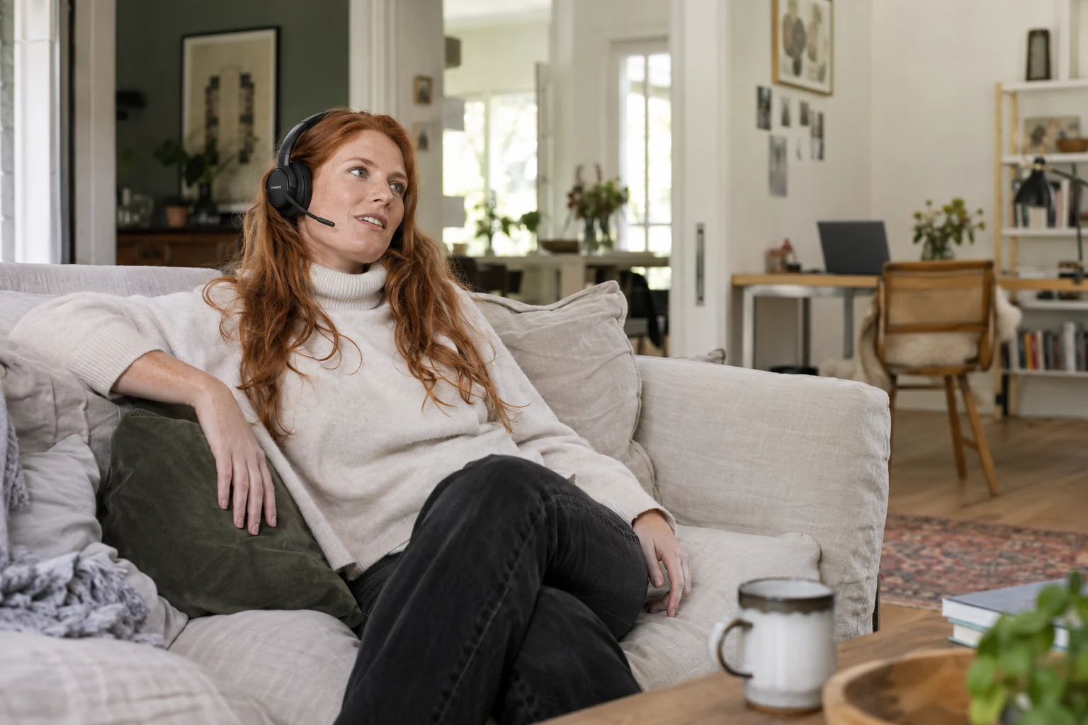 KI-generierte Szene: Eine rothaarige Frau im Rollkragen diktiert entspannt auf dem Sofa über ein Headset; der Laptop steht hinten rechts.